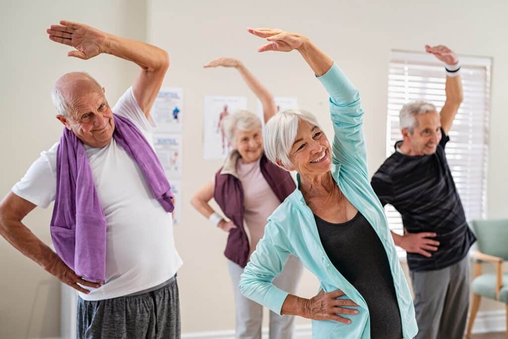 Group of older adults doing side-stretching exercises in a bright room, smiling as they reach one arm over their heads.